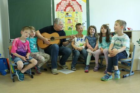 Eine Frau spielt Gitarre für eine Gruppe von Kindern in der Kita in einem Raum mit einer grünen Tafel im Hintergrund, auf der ein Plakat hängt.